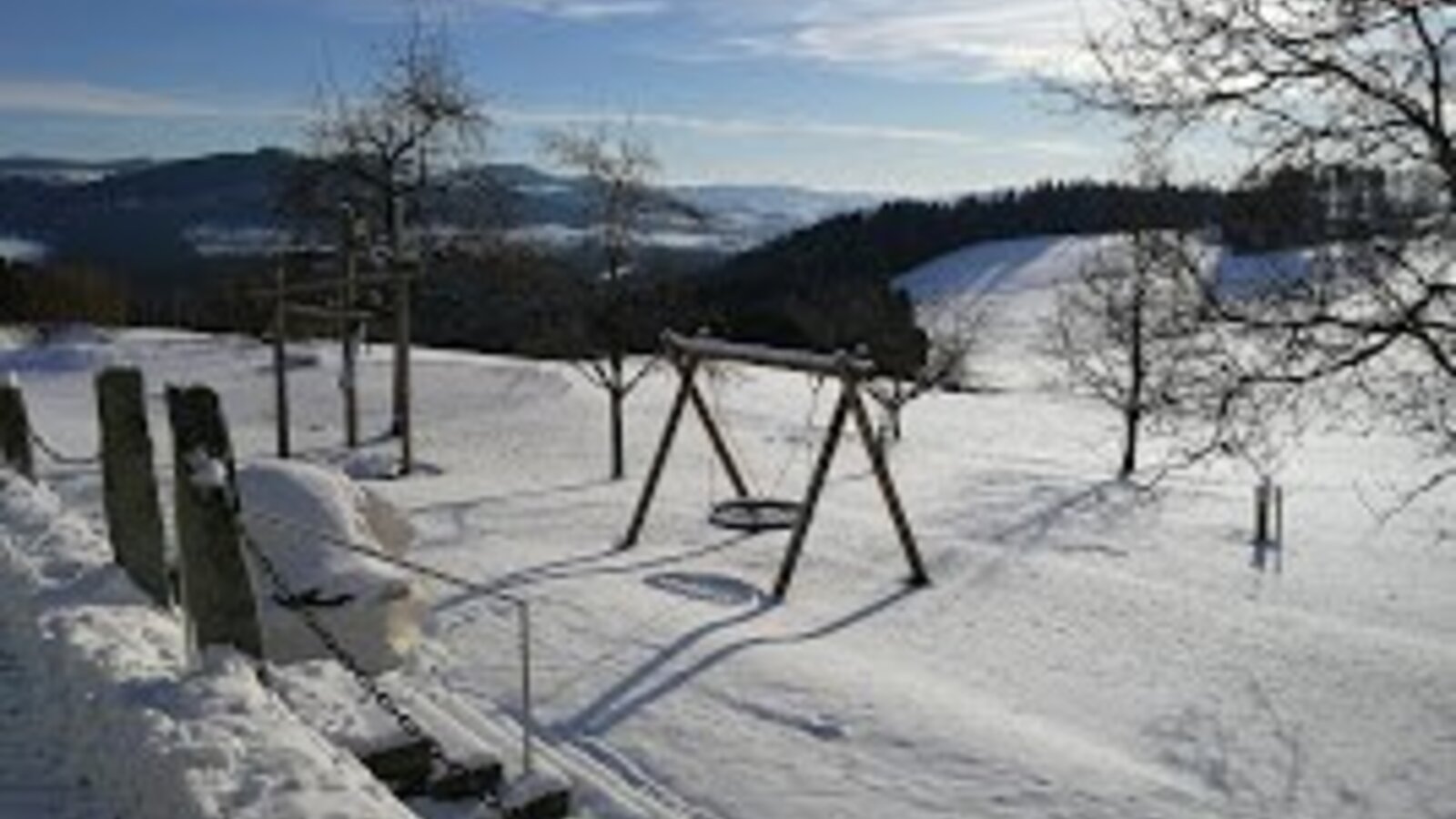 Der verschneite Spielbereich des Bauernhofs mit Schaukel und Sitzbank.