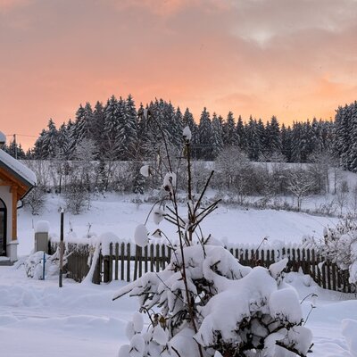 Winterliche Landschaft des Bauernhofs mit einer kleinen Kapelle, verschneitem Wald und einem Himmel in warmen Farbtönen.