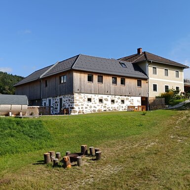 Außenansicht des Bauernhofs mit seinen zwei Gebäudeteilen, einer Fasssauna, einer Feuerstelle mit Holzstumpf-Sitzgelegenheiten und einer weitläufigen Grünfläche, umgeben von Wald.