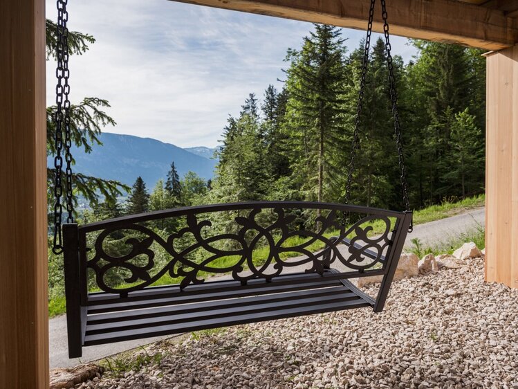 Metallene Schaukelbank auf der überdachten Terrasse des Hofs mit Blick auf Wald und Berge.