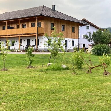 Außenansicht des Bauernhofs mit Rasenfläche, Terrassen mit Sitzgelegenheiten im Erdgeschoss und einem Balkon im Obergeschoss.