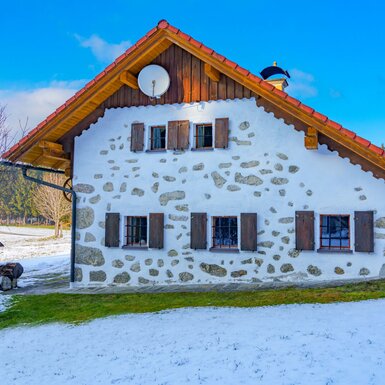 Außenansicht des Bauernhofs mit Fassade aus Stein und weißem Putz, Holzläden und verschneitem Gartenbereich.