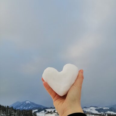 Hand hält ein Schneeherz vor der winterlichen Berglandschaft rund um den Bauernhof.