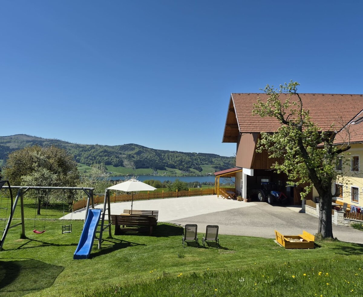 Außenansicht des Bauernhofs mit Garten, Kinderspielplatz, Sitzgelegenheiten und Blick auf den See und die Berge.