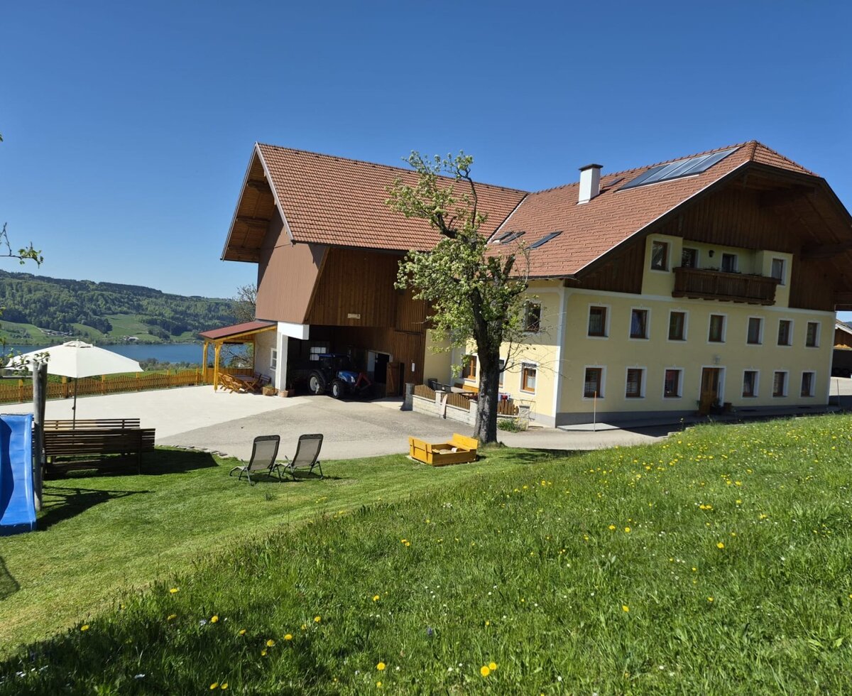 Der Bauernhof mit Gartenbereich, Rutsche, Sonnenliegen und überdachtem Sitzbereich, mit Blick auf den See und die Berge.