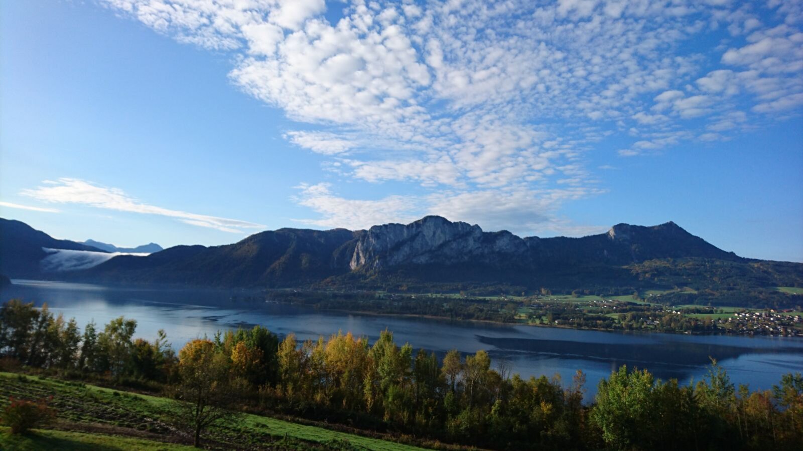 Blick auf den See und die umliegenden Berge mit herbstlicher Vegetation im Vordergrund vom Bauernhof aus.