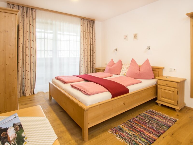 Doppelzimmer im Bauernhof mit Holzdoppelbett, zwei Nachttischen, Holzschrank und einem Fenster mit Vorhängen.