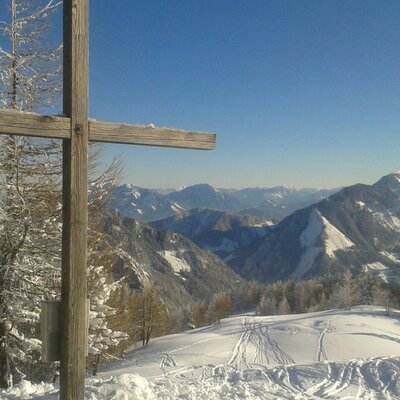 Die verschneite Berglandschaft in der Umgebung des Bauernhofs zeigt ein Gipfelkreuz, frostbedeckte Bäume und Skispuren unter blauem Himmel.