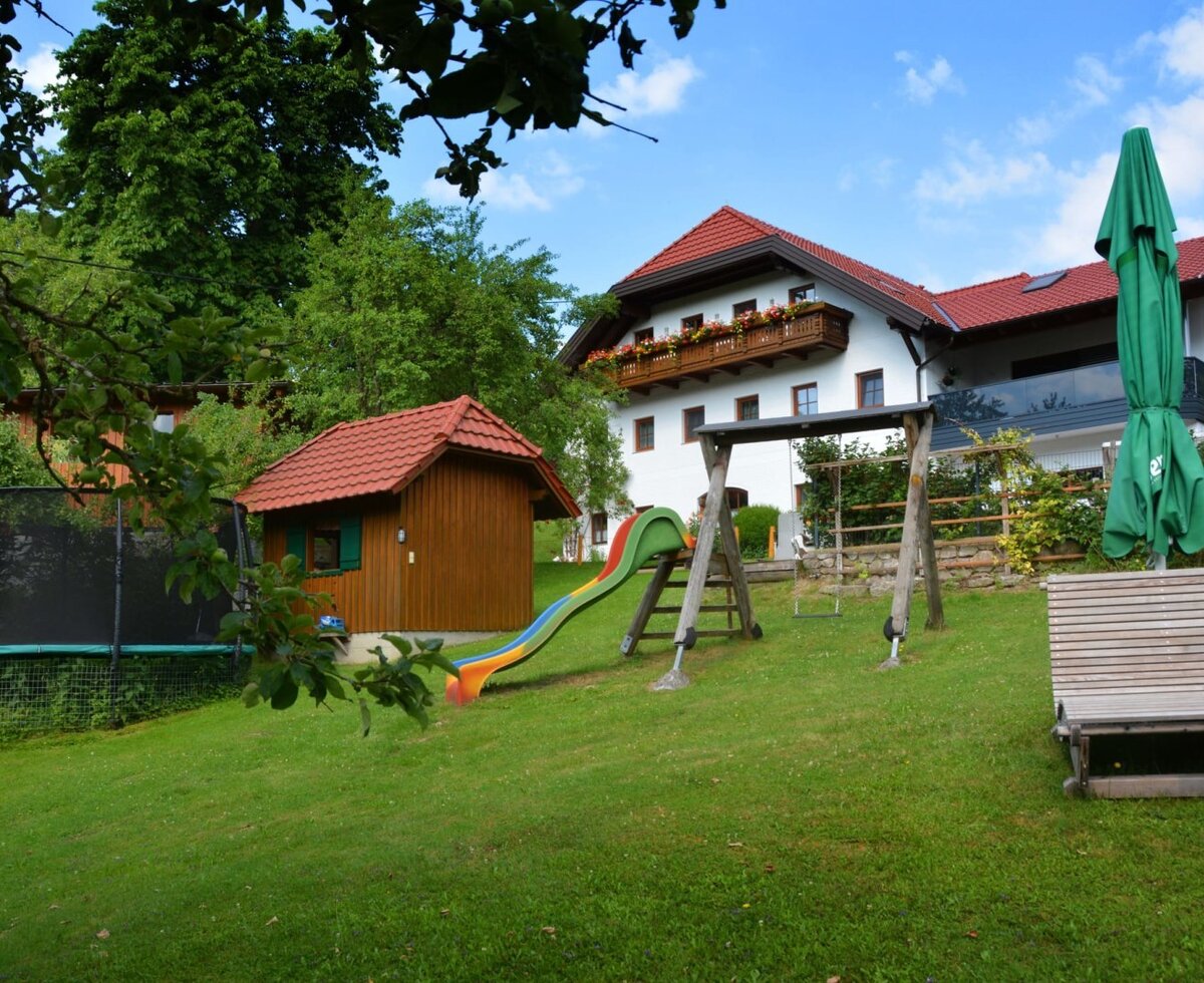 Der Außenbereich des Bauernhofs mit einem Spielplatz inklusive Rutsche, Schaukel und Trampolin, einem Spielhaus und einer Holzliege auf der Rasenfläche.
