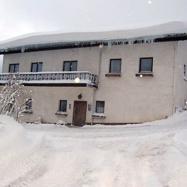 Außenansicht des Bauernhofs im Winter, mit Schnee auf dem Dach, Eiszapfen, Balkon und schneebedecktem Vorplatz.