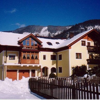 Der Bauernhof mit schneebedecktem Dach und Holzbalkonen vor einer winterlichen Berglandschaft, mit einem Zaun im Vordergrund.