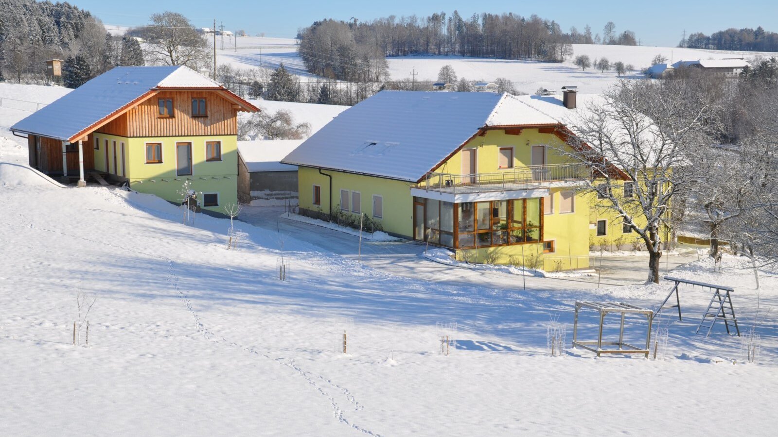 Die Gebäude des Bauernhofs im Winter, mit dem Wintergarten und einem Schaukelgestell auf dem verschneiten Spielplatz.