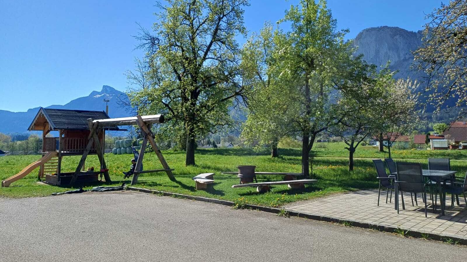 Der Kinderspielplatz mit Spielhaus und Schaukel sowie die Sitzgelegenheiten im Freien auf dem Bauernhof, mit Blick auf die umliegenden Wiesen und Berge.