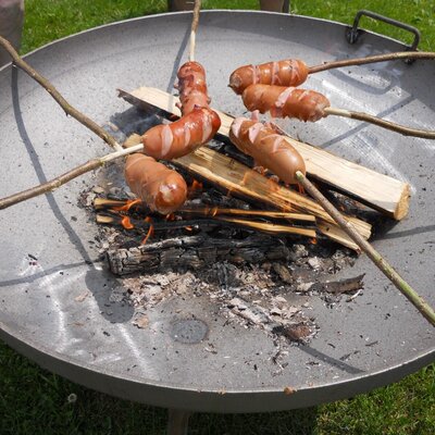 Knackwürste auf Grillstäben über einem offenen Feuer einer Grillschale.
