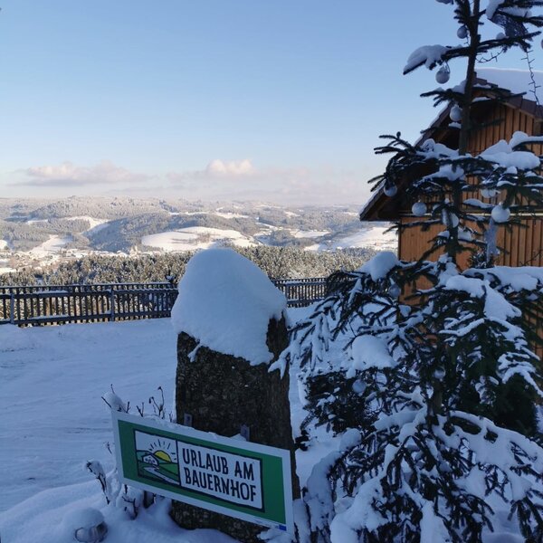 Hinweisschild "Urlaub am Bauernhof" im Schnee mit Panoramablick über die winterliche Landschaft vom Bauernhof.