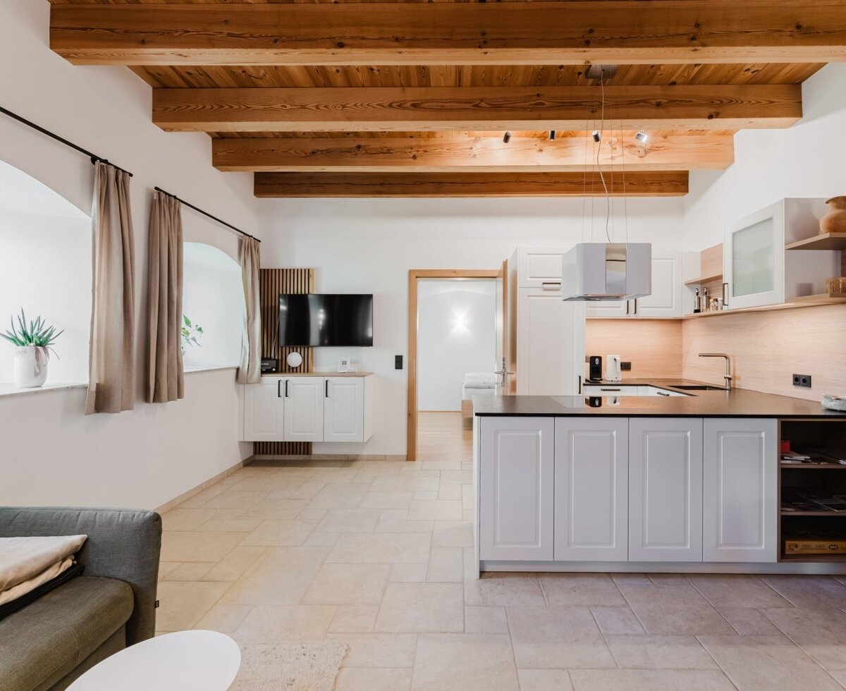 Offener Wohn- und Küchenbereich der Ferienwohnung mit moderner Kochinsel, Sofa, Fernseher und sichtbaren Holzbalken.