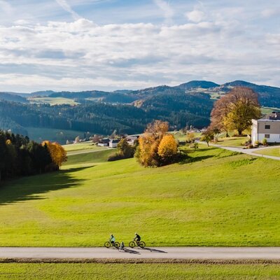 Zwei Radfahrer auf einer Straße vor der hügeligen Mühlviertler Landschaft, mit den Gebäuden des Bauernhofs im Hintergrund.
