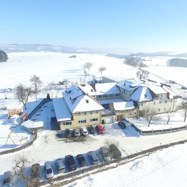 Der Bauernhof mit seinen Gebäuden, umgeben von verschneiter Winterlandschaft, einem Kinderspielplatz und Parkmöglichkeiten.