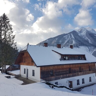 Außenansicht des Bauernhofs mit schneebedecktem Dach und Holzfassade vor einer Bergkulisse.