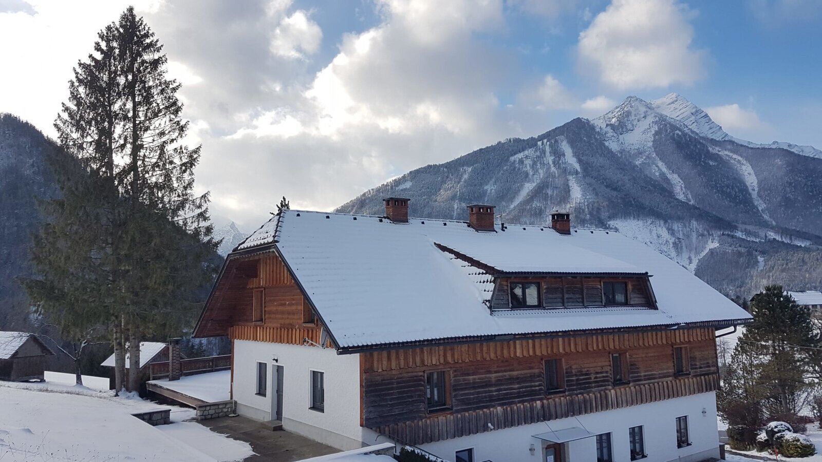 Außenansicht des Bauernhofs mit schneebedecktem Dach und Holzfassade vor einer Bergkulisse.