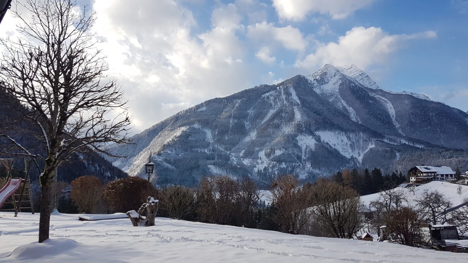 Der verschneite Gartenbereich des Bauernhofs mit sichtbarem Spielplatz und Bergen im Hintergrund.