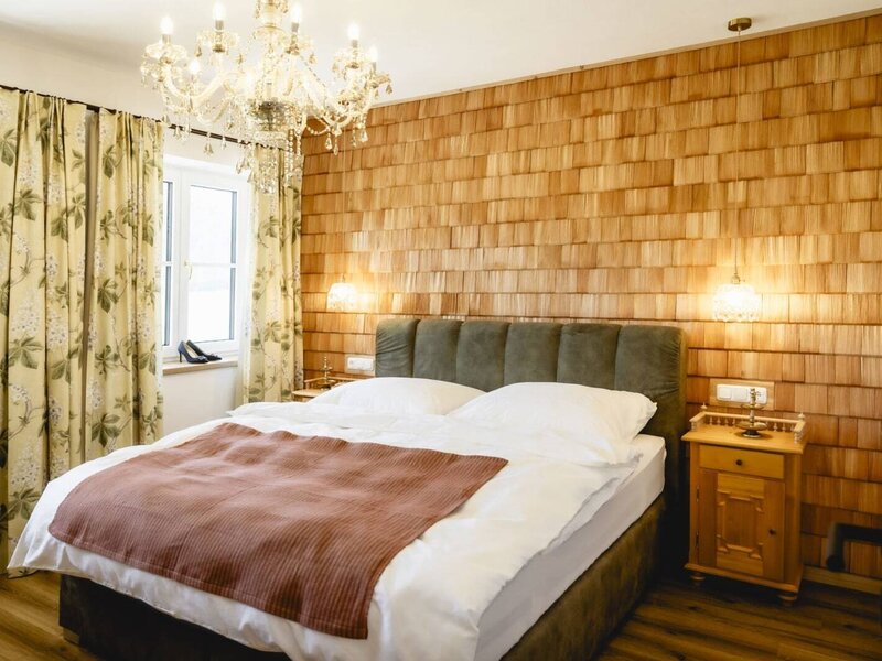 Schlafzimmer im Appartement mit Doppelbett, Holzschindelwand, Fenstern mit floralen Vorhängen und Kronleuchter.
