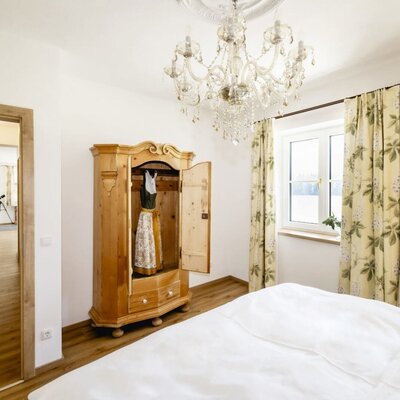Schlafzimmer im Appartement mit Bett, traditionellem Holzschrank mit Dirndl, Kronleuchter und Fenstern.
