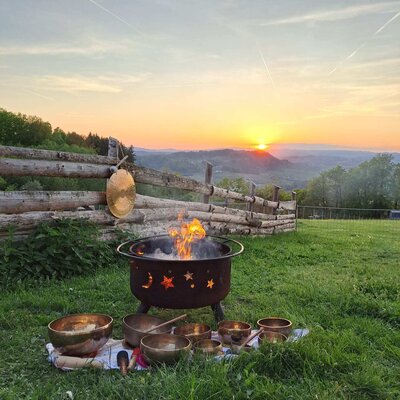 Eine gemütliche Feuerschale und Klangschalen auf der Wiese vor einem stimmungsvollen Sonnenuntergang.