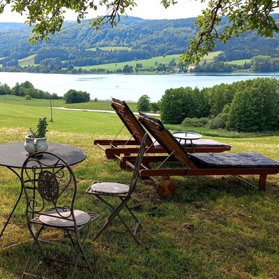 Außenbereich des Bauernhofs mit Tisch, Stühlen und Liegestühlen unter einem Baum, mit Blick auf den See und die umliegenden Hügel.