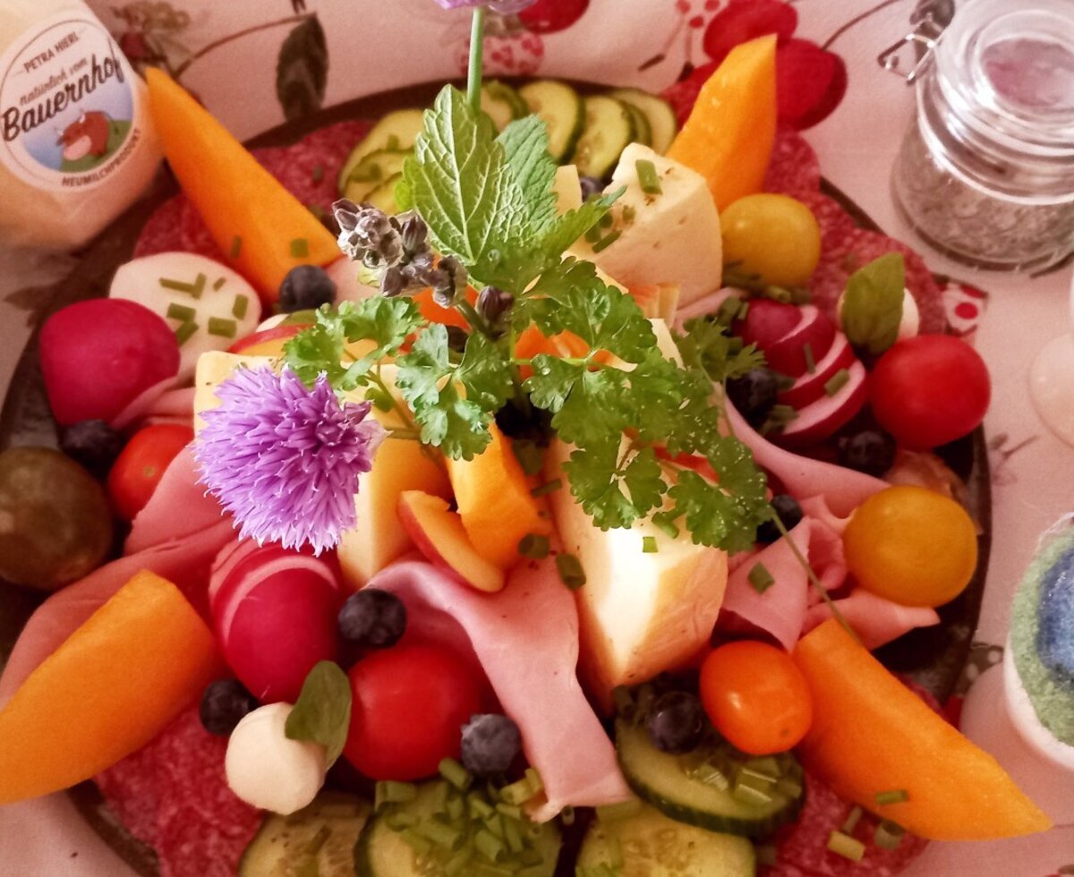 Eine Frühstücksplatte mit Käse, Wurst, frischem Gemüse, Obst und Kräutern, ergänzt durch Eier und weitere Bauernhofprodukte.