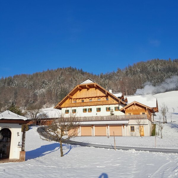 Der Bauernhof im Winter mit schneebedeckter Landschaft, Wald im Hintergrund und einem kleinen Schrein im Vordergrund.