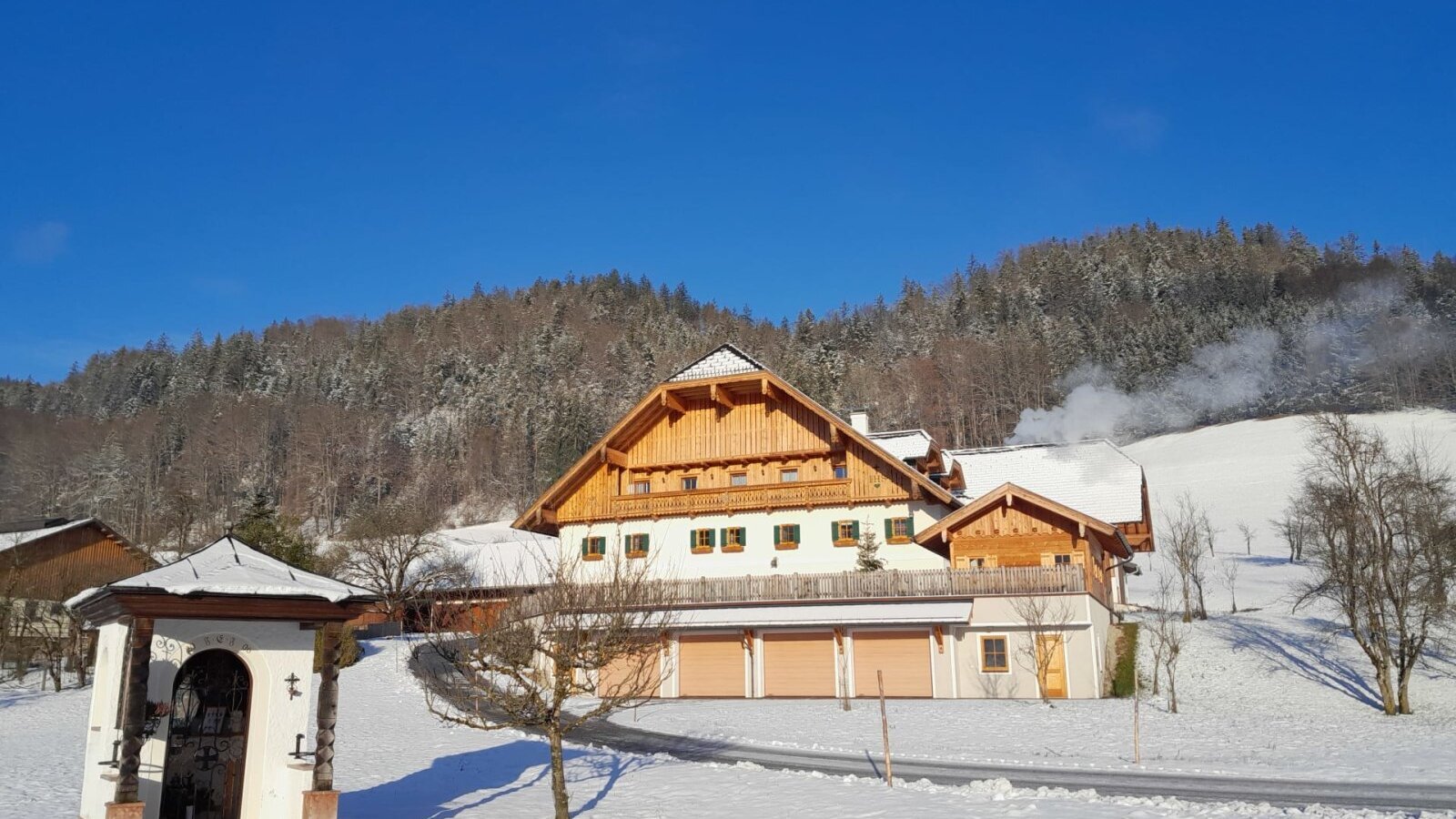 Der Bauernhof im Winter mit schneebedeckter Landschaft, Wald im Hintergrund und einem kleinen Schrein im Vordergrund.