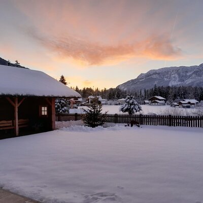 Winterlandschaft des Bauernhofs mit schneebedecktem Garten, Holzpavillon und Bergen im Hintergrund.
