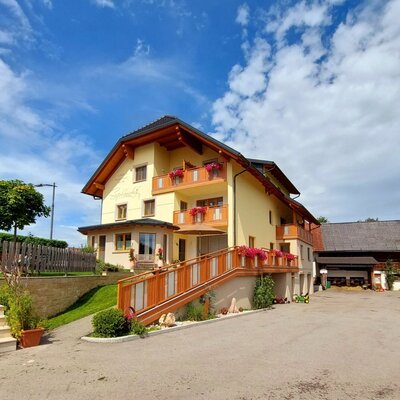 Das Hauptgebäude des Bauernhofs mit gelber Fassade, Holzbalkonen, einer Zugangstreppe mit Blumen und einem Kinderspielplatz.