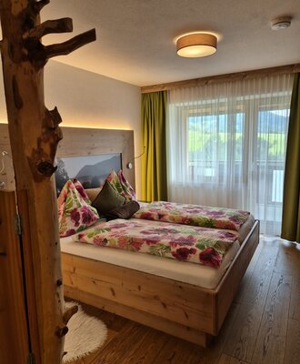 Doppelzimmer im Bauernhof mit naturbelassenen Holzelementen, einem Holzbett mit floraler Bettwäsche und Fensterblick auf die grüne Landschaft.