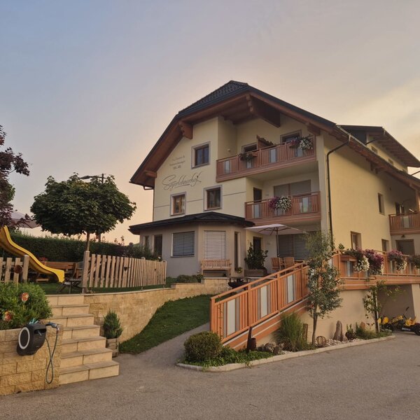 Der Bauernhof mit Balkonen, einer Terrasse mit Sonnenschirm und einem Kinderspielplatz mit Rutsche.