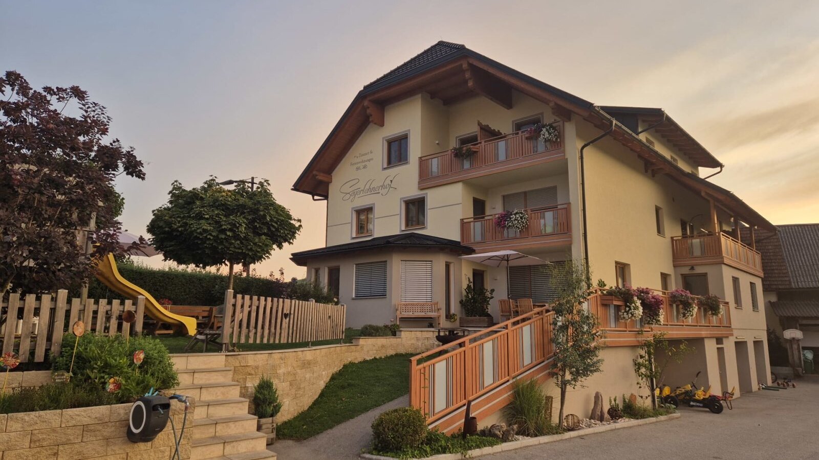 Der Bauernhof mit Balkonen, einer Terrasse mit Sonnenschirm und einem Kinderspielplatz mit Rutsche.