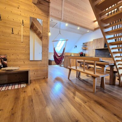 Der Innenbereich des Bauernhofs mit Holzboden, Holzwänden, einer Sitzbank, einem Esstisch mit Bänken, einer Küche, einer Hängematte und einer Holztreppe.