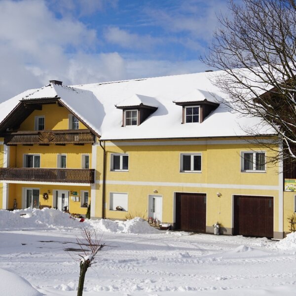 Der Bauernhof mit gelber Fassade, Balkon, schneebedecktem Dach und großen Toren im Erdgeschoss.