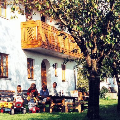 Außenansicht des Bauernhofs mit einem Balkon und einem Gartenbereich mit Holztisch und Spielmöglichkeiten für Kinder.