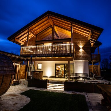 Das Traunsee-Chalet am Abend mit beleuchtetem Balkon, Hot-Pot, Fasssauna und einer Terrasse mit Sitzgelegenheiten.