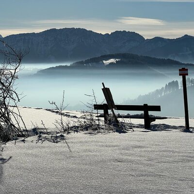 Eine Sitzbank im Schnee mit Blick über das nebelverhangene Tal und die umliegenden Berge vom Bauernhof.