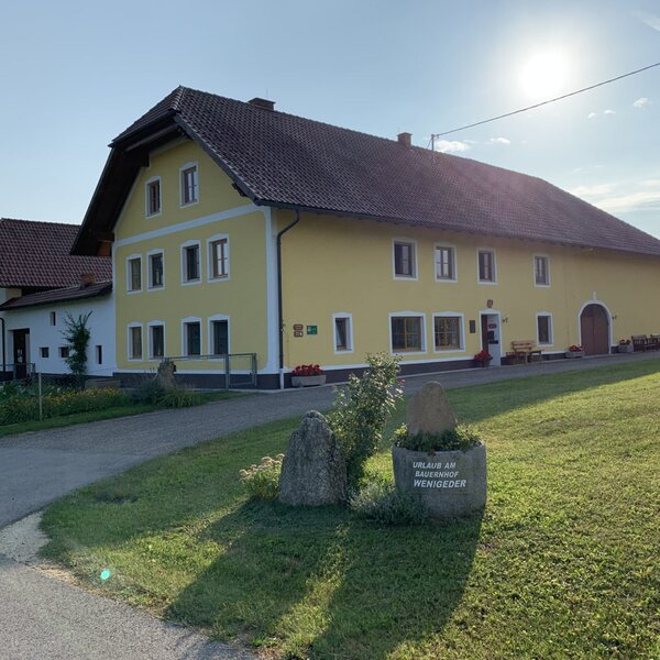 Der einladende Bauernhof präsentiert sich mit einem gepflegten grünen Rasen und einer freundlichen gelben Fassade im Sonnenschein.