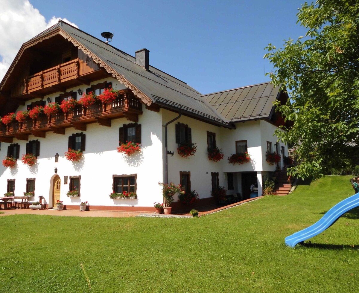Die Außenansicht des Bauernhofs mit blühenden Holzbalkonen, einer Sitzgelegenheit im Freien und einer Rasenfläche mit Kinderrutsche.
