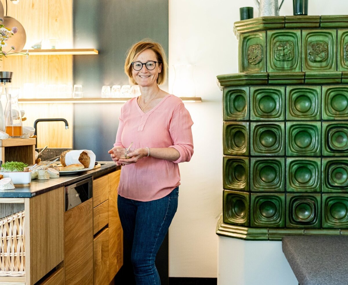 neue Stube - Die Gastgeberin Sabine neben dem Frühstücksbuffet mit frischen Speisen und einem traditionellen Kachelofen.