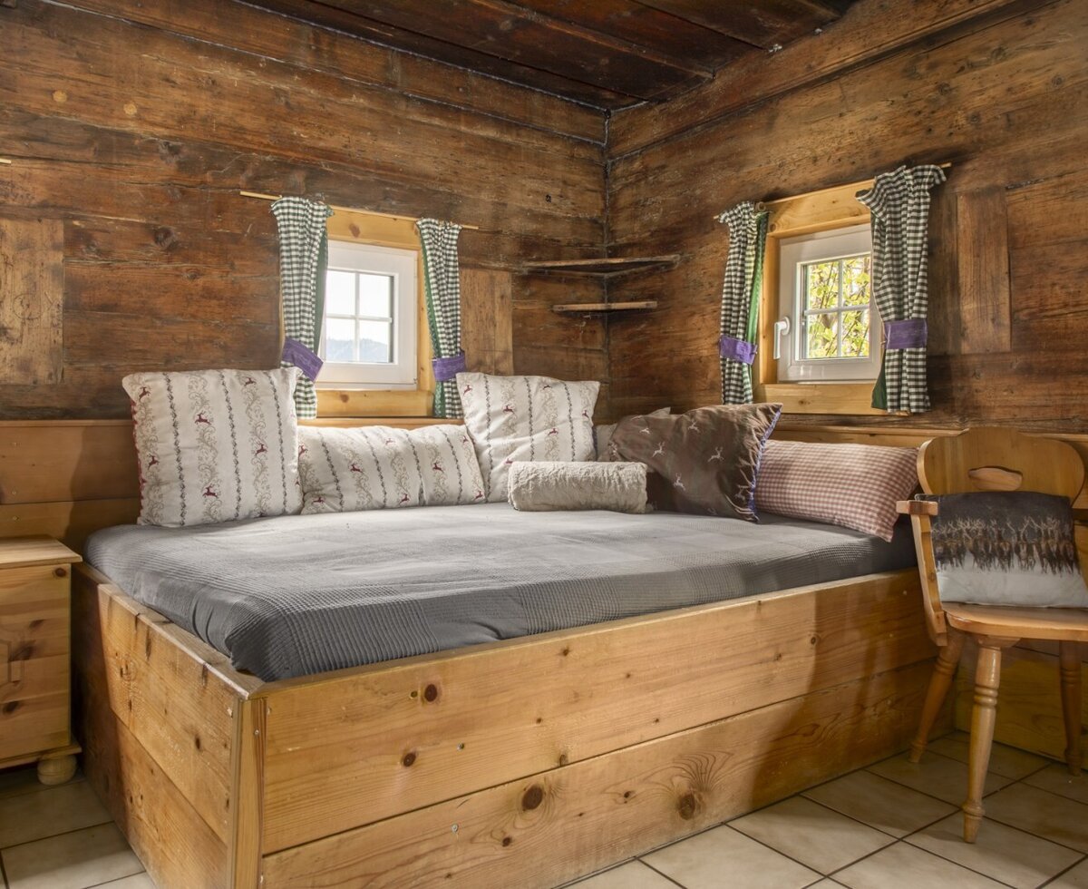 Schlafbereich in der Almhütte mit einem Holzbett, Fenstern und traditionellen karierten Vorhängen.