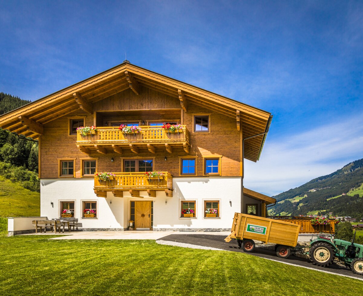 Der Bauernhof mit Holzfassade, Balkonen mit Blumenkästen und einer Terrasse mit Sitzgelegenheiten im Garten, umgeben von grünen Bergen.