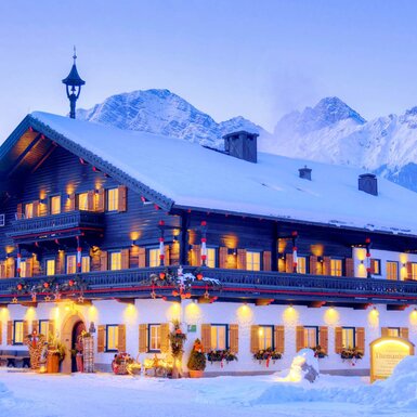 Die Außenansicht des Bauernhofs, umgeben von Schnee, mit erleuchteten Fenstern und Blick auf die verschneiten Berge.