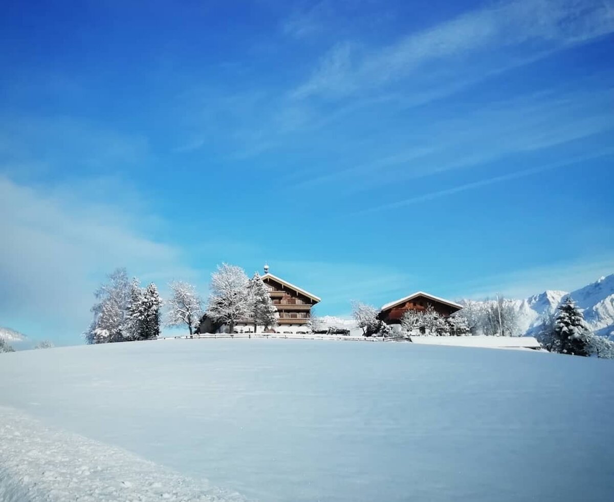 Der Bauernhof in einer schneebedeckten Winterlandschaft, mit traditionellen Holzhäusern und Blick auf ferne Berge.