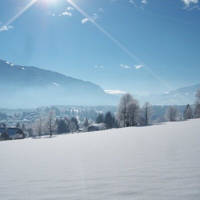 Blick vom Bauernhof auf die verschneite Winterlandschaft mit Feldern, Bergen und einem Dorf.